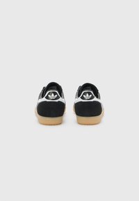 Des baskets en suede noir avec des accents en cuir blanc, dotées d'une semelle en caoutchouc texturé et d'un logo noir reconnaissable sur le talon.