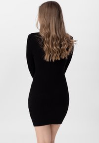 Robe noire en maille côtelée avec manches longues, silhouette ajustée et col montant, mettant en valeur un tissu texturé et une longueur au-dessus du genou.