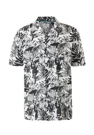 Personne portant une chemise à manches courtes à imprimé tropical noir et blanc, un short beige, des chaussettes noires et des sandales noires, debout les mains dans le dos.