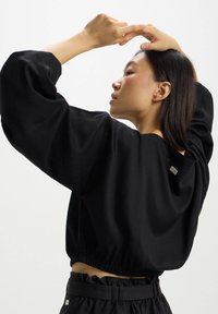 Blusa nera corta con maniche lunghe e voluminosi, dotata di un elastico in vita e un piccolo tag con logo sul retro. Tessuto dalla texture liscia.