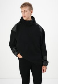 Μαύρο oversized hoodie με απαλή υφή, που διαθέτει ματ μαύρες λεπτομέρειες στους ώμους και ψηλό λαιμό. Συνδυάζεται με μαύρο παντελόνι.