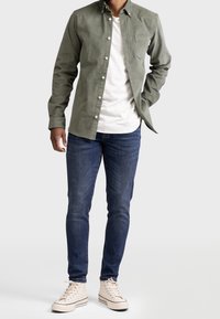 Camisa verde de manga larga con un bolsillo en el pecho, combinada con jeans azules y zapatillas altas beige. La camisa presenta una tela texturizada.