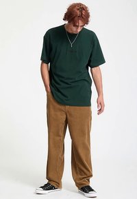Camiseta verde oscuro de manga corta y cuello redondo, combinada con pantalones de pana marrones. Zapatillas negras con suelas blancas.