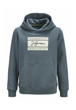 Personne portant une veste en polaire à motifs, un sweat à capuche bleu avec un logo, un jean ample à lavage léger et des baskets blanches, debout devant un fond uni.