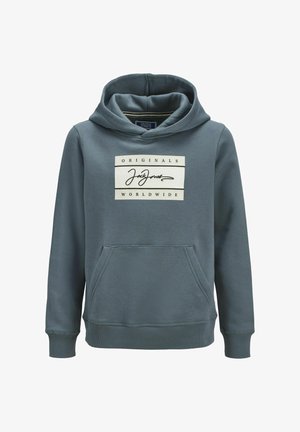 Personne portant une veste en polaire à motifs, un sweat à capuche bleu avec un logo, un jean ample à lavage léger et des baskets blanches, debout devant un fond uni.