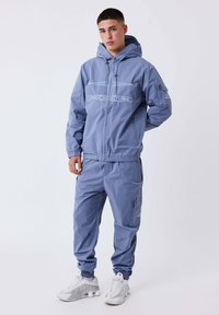 Giovane uomo che indossa una tuta blu con cappuccio e sneakers bianche, in piedi con una mano in tasca su uno sfondo neutro.