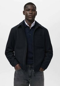 Chaqueta navy con cremallera y cuello, bolsillo frontal y tejido texturizado. Llevada sobre un suéter oscuro y jeans de mezclilla gris.