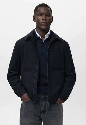 Chaqueta navy con cremallera y cuello, bolsillo frontal y tejido texturizado. Llevada sobre un suéter oscuro y jeans de mezclilla gris.