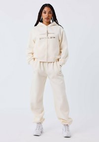 Sudadera con cremallera color crema con un logo y pantalones de chándal a juego. Hecha de tela suave, presenta puños acanalados y un ajuste relajado.