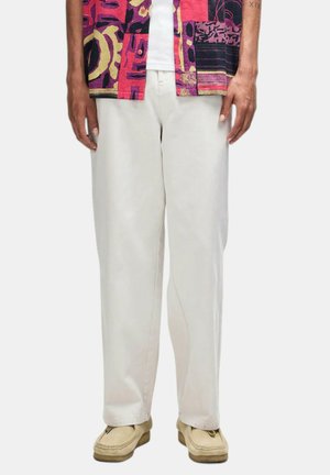 Persona che indossa pantaloni bianchi larghi, mocassini beige e una camicia colorata rossa, rosa e gialla con motivi sopra una canottiera bianca.