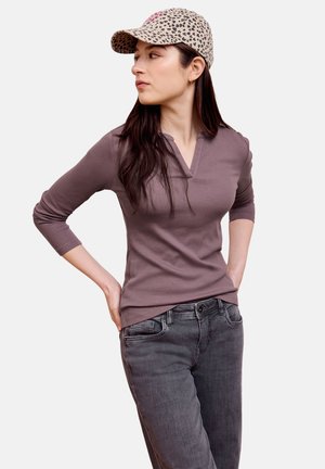 Mujer con un top de punto marrón, jeans gris oscuro y una gorra beige con estampado de leopardo, de pie con las manos en las caderas, mirando hacia la izquierda.