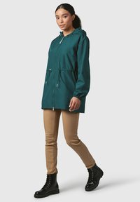Giacca leggera di color teal con cappuccio, polsini elasticizzati e vita regolabile con cordino. Indossata con pantaloni color sabbia e stivaletti neri con lacci.