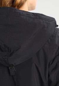 Czarna kurtka z kapturem, z detalami w przeszyciach i regulowanym sznurkiem. Materiał wydaje się gładki i lekki, idealny do użytku na świeżym powietrzu.