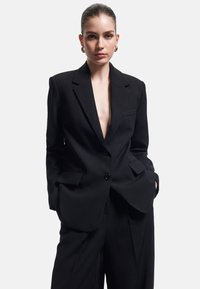 Zwart op maat gemaakte blazer met een diepe V-hals, een enkele knoopsluiting en twee voorzakken. Een paar zwarte wijde broek is de perfecte aanvulling op de outfit.