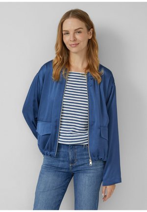 Vrouw in een blauwe ritsjack over een wit en blauw gestreept shirt en blauwe jeans, staande tegen een effen lichte achtergrond.