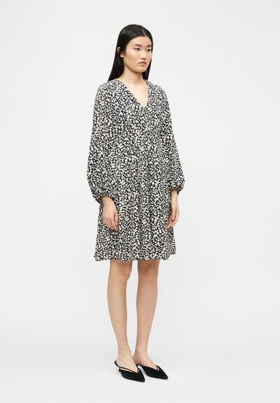 Γυναίκα που στέκεται, φορώντας φόρεμα μήκους μέχρι τα γόνατα με άσπρα και μαύρα λουλούδια, με μακριά φουσκωτά μανίκια και μαύρα μυτερά παπούτσια slingback.