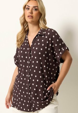 Blonde vrouw die een losse bruine korte mouwen blouse met witte stippen en een witte broek draagt, poseert met één hand in de zak.