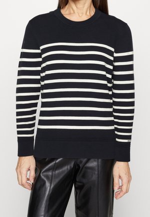 Person trägt einen schwarz-weißen, horizontal gestreiften Pullover mit langen Ärmeln und schwarze Lederhose, steht vor einem einfarbigen Hintergrund.