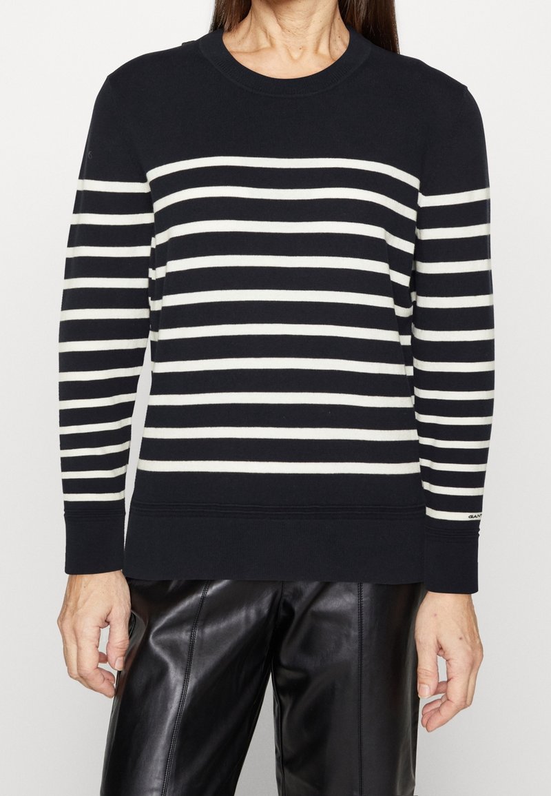 Personne portant un pull à manches longues à rayures horizontales noires et blanches et un pantalon en cuir noir, debout devant un fond uni.