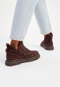 Bottes chevilles en daim marron avec un design à lacets, surface texturée et semelle en caoutchouc. Présente une languette arrière et un discret détail de couture.