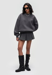 Dunkelgrauer übergroßer Sweatshirt mit Logo, grauer plissierter Minirock und schwarze kniehohe Stiefel mit Schnallen-Details. Model trägt Sonnenbrille.