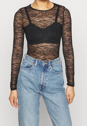 Persona con una blusa de encaje floral negra de manga larga, un bralette negro debajo y unos vaqueros de mezclilla azul claro de talle alto.