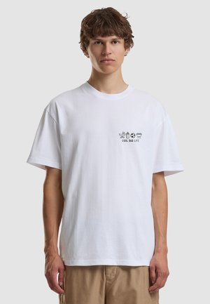 Young man wearing a white T-shirt with "COOL DAD LIFE" and icons of stroller, baby bottle, soccer ball, and grill on the chest.