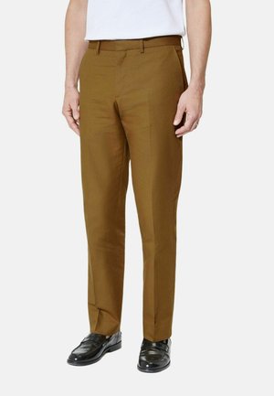 Mężczyzna ubrany w brązowe, dopasowane spodnie, białą koszulę włożoną do środka i czarne skórzane eleganckie buty, stojący na jednolitym tle.