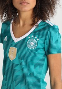 Teal jalkapalloasu, jossa on geometrisia kuvioita, valkoinen kaulus, Adidas-logo, Saksan jalkapalloliiton merkki ja FIFA 2014 maailmanmestaruuskilpailujen patch.