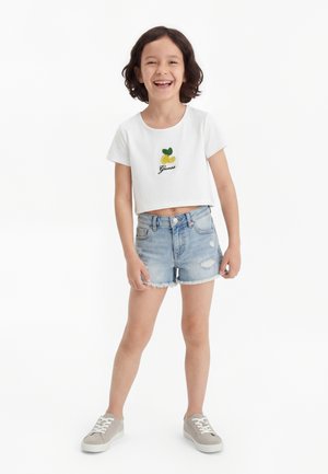 Χαμογελαστό παιδί με σκούρα μαλλιά φορώντας λευκό κοντομάνικο μπλουζάκι με κέντημα λεμονιού, ανοιχτόχρωμα τζιν σορτς και γκρι αθλητικά παπούτσια, στέκεται σε λευκό φόντο.