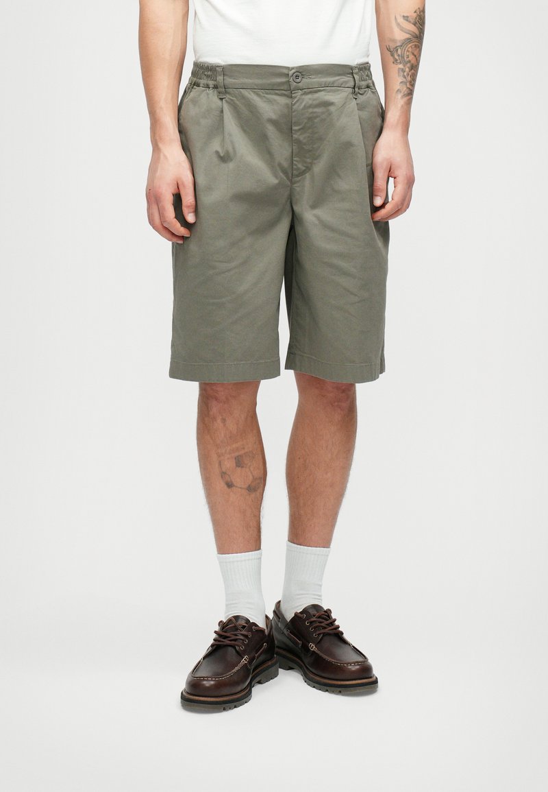 Homme portant un short vert olive, des chaussettes blanches et des chaussures à lacets marron foncé, se tenant devant un fond blanc uni.