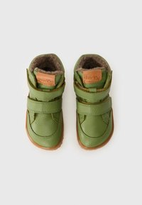 Botas de cuero verde con dos correas de velcro, suelas de goma marrón y revestimiento de suave piel sintética. Presenta un parche rectangular de logotipo beige en el lateral.