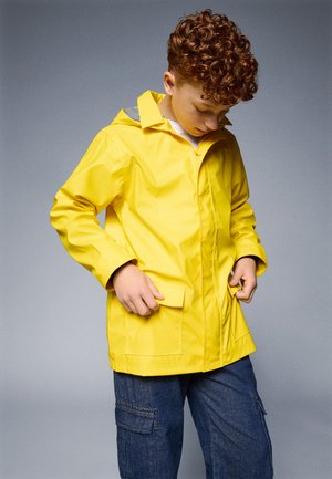 Giacca antipioggia gialla e impermeabile con zip frontale, due tasche a patta e fodera a righe nel cappuccio, indossata sopra pantaloni cargo blu.