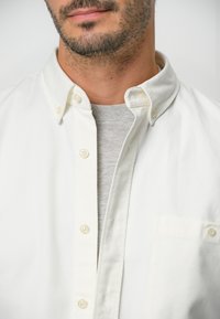 Witte gekraagde overhemd met een borstzak, gelaagd over een lichtgrijze ronde hals t-shirt; heeft lichte knopen en een gladde textuur.