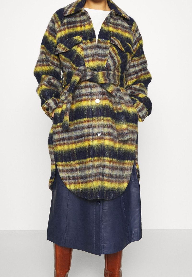 Femme portant un manteau en laine ceinturé à rayures jaunes, noires et grises, par-dessus une jupe midi en cuir bleu marine et des bottes marron montant jusqu'aux genoux.