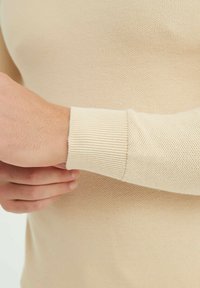 Pull en tricot beige avec une finition texturée, des poignets côtelés et une coupe ajustée, mettant en valeur un tissu doux et léger.