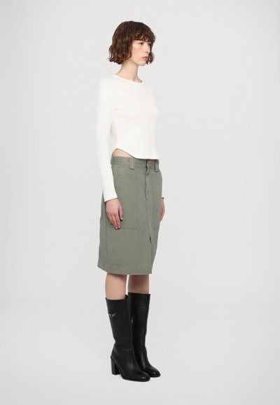 White long-sleeve top, cropped design. Olive green skirt with front pockets and center slit. Black knee-high boots with block heels.