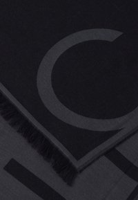 Fekete és sötétszürke sál rojtos szegéllyel, geometrikus betűkkel és mintákkal, puha anyagból készült, mely kiemeli a textúrát és a dizájnt.