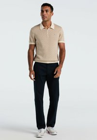 Beige polo shirt met een patroon, korte mouwen en een crèmekleurige kraag, gecombineerd met donkerblauwe pantalon en witte sneakers.