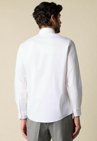 Witte langslobige shirt met hoge kraag, gladde textuur en subtiele ronde zoom, gecombineerd met grijze broek met streepdessin.