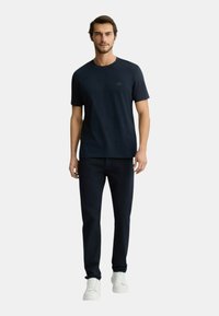 Maglietta in cotone blu navy con colletto rotondo e maniche corte, abbinata a pantaloni scuri e sneakers bianche, si trova davanti a uno sfondo neutro.