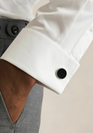 Left wrist with dark skin wears a white dress shirt cuff fastened by a black and silver cufflink, hand in gray trousers pocket.