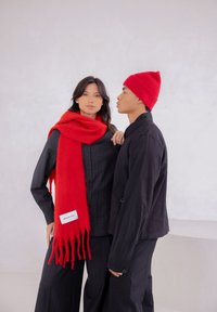 Rode wollen sjaal met gefranjerde uiteinden, gedragen over een zwart overhemd met krijtstrepen. Zwarte jas op model ernaast, met een bijpassende rode gebreide muts.