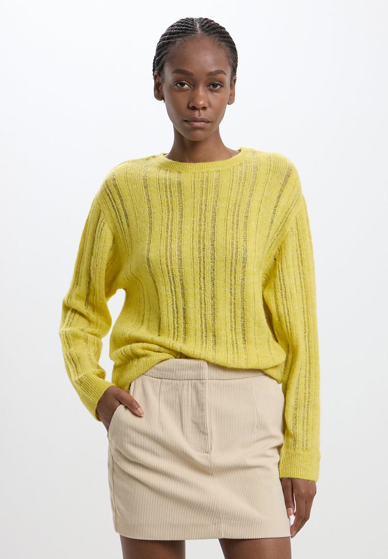 Maglione giallo a maglia con costine verticali, caratterizzato da maniche lunghe e collo rotondo, abbinato a una gonna beige con texture.