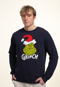 Námořnická modrá mikina se zobrazením Grinche v červené a bílé vánoční čepici a slovem "GRINCH" v tučných bílých písmenech.