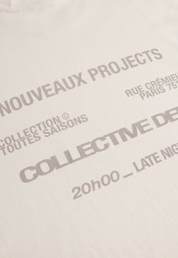 Weißes Baumwoll-T-Shirt mit auffälligem grauem Text. Der Text umfasst "NOUVEAUX PROJECTS", "COLLECTIVE DE" und Veranstaltungsdetails. Sanfte Textur.