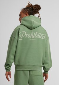 Sweatshirt vert surdimensionné avec une finition texturée, arborant le logo "Interdit" dans le dos et un design de poche kangourou.