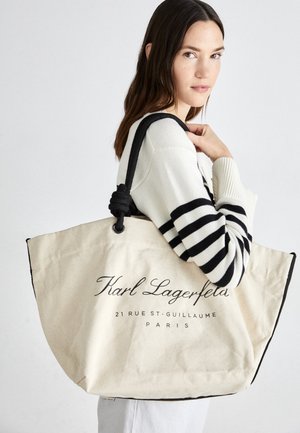 Vrouw in gestreepte trui met een grote beige tas met zwarte hengsels en de tekst "Karl Lagerfeld Paris".