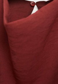 Textilie v rezavé barvě s jemnou strukturou, která má částečný límec a detail knoflíku na výstřihu.