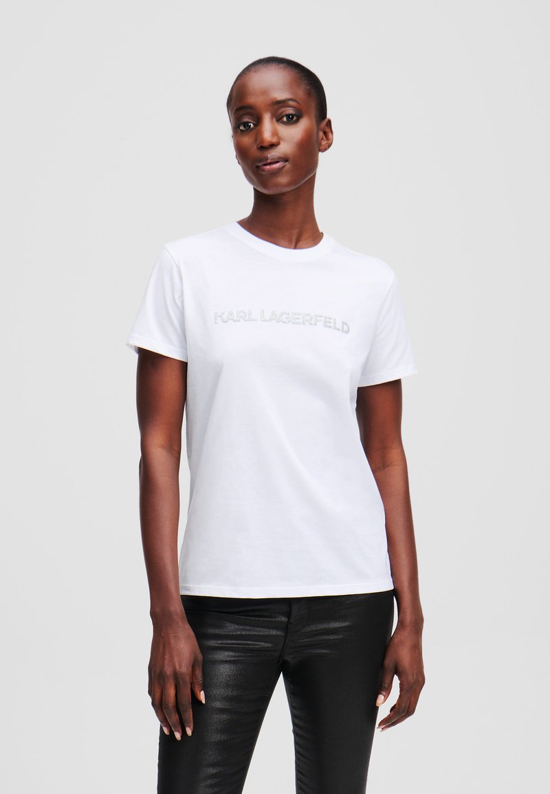 Weißes Baumwoll-T-Shirt mit kurzen Ärmeln, das ein silbernes "KARL LAGERFELD"-Logo über der Brust trägt. Runder Ausschnitt, klassischer Schnitt.